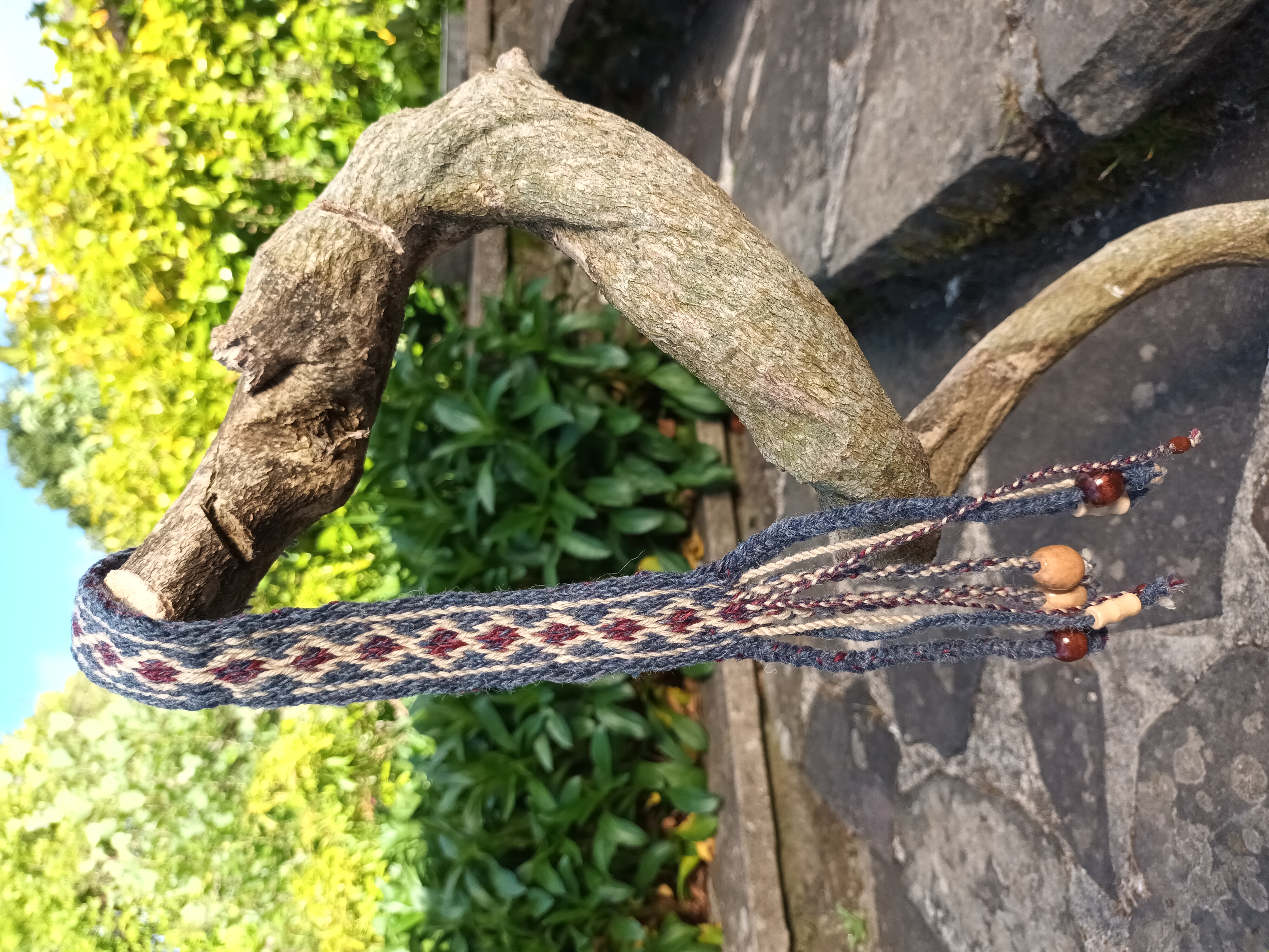 Blue, cream and burgundy card woven band hanging from a curved branch with wooden beads